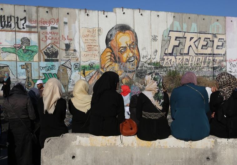 Palestinian women sit near a section of Israel's separation barrier at Qalandia checkpoint in front of a painted mural of Marwan Barghouti
